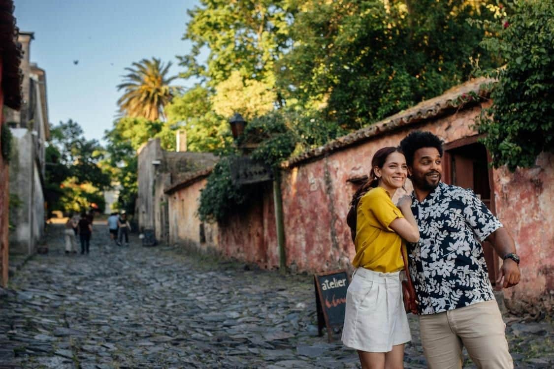 A smiling couple poses outdoors near a rustic stone path and a wall with greenery, under a bright and sunny sky.