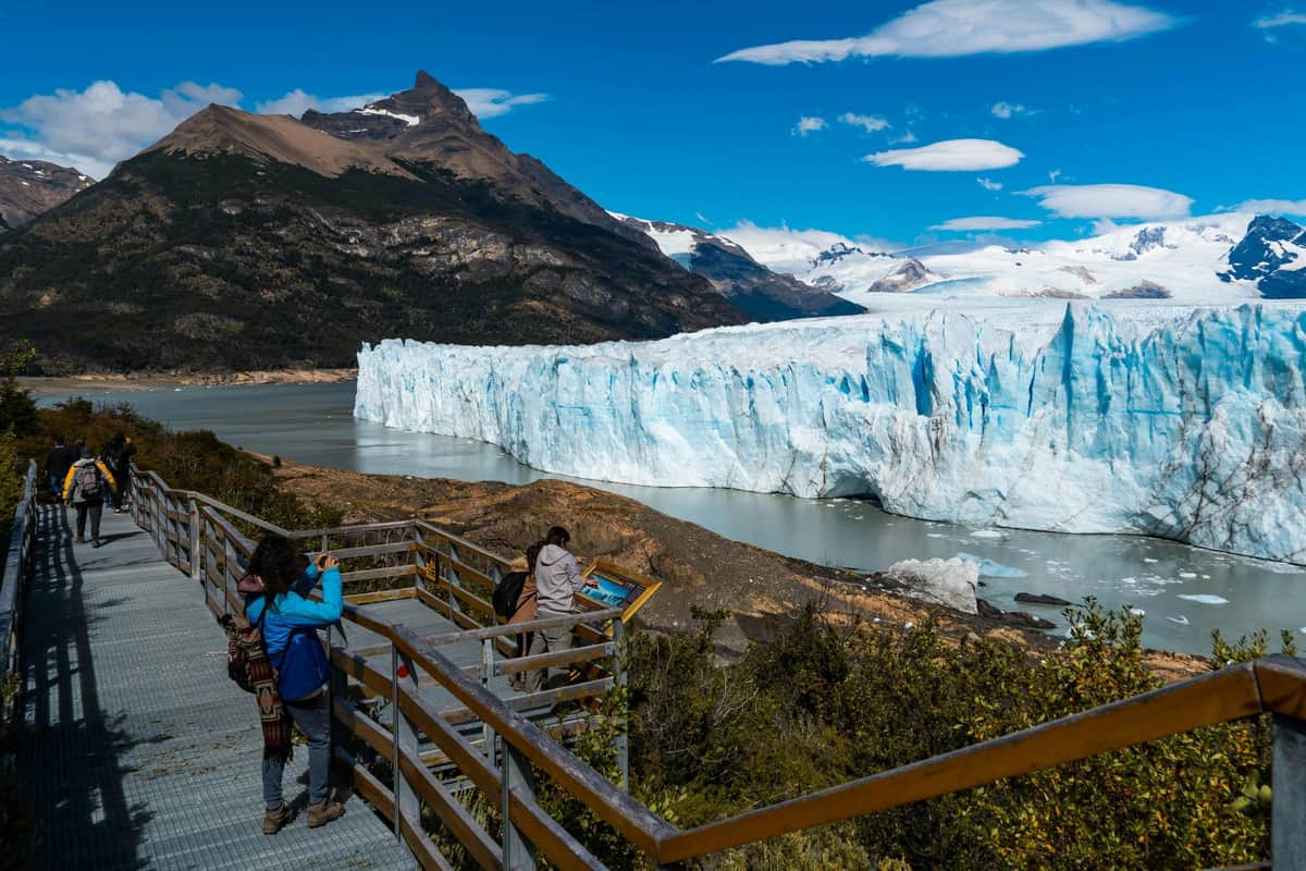 Tourists on a wooden walkway viewing a large glacier surrounded by mountains and water under a partly cloudy sky.