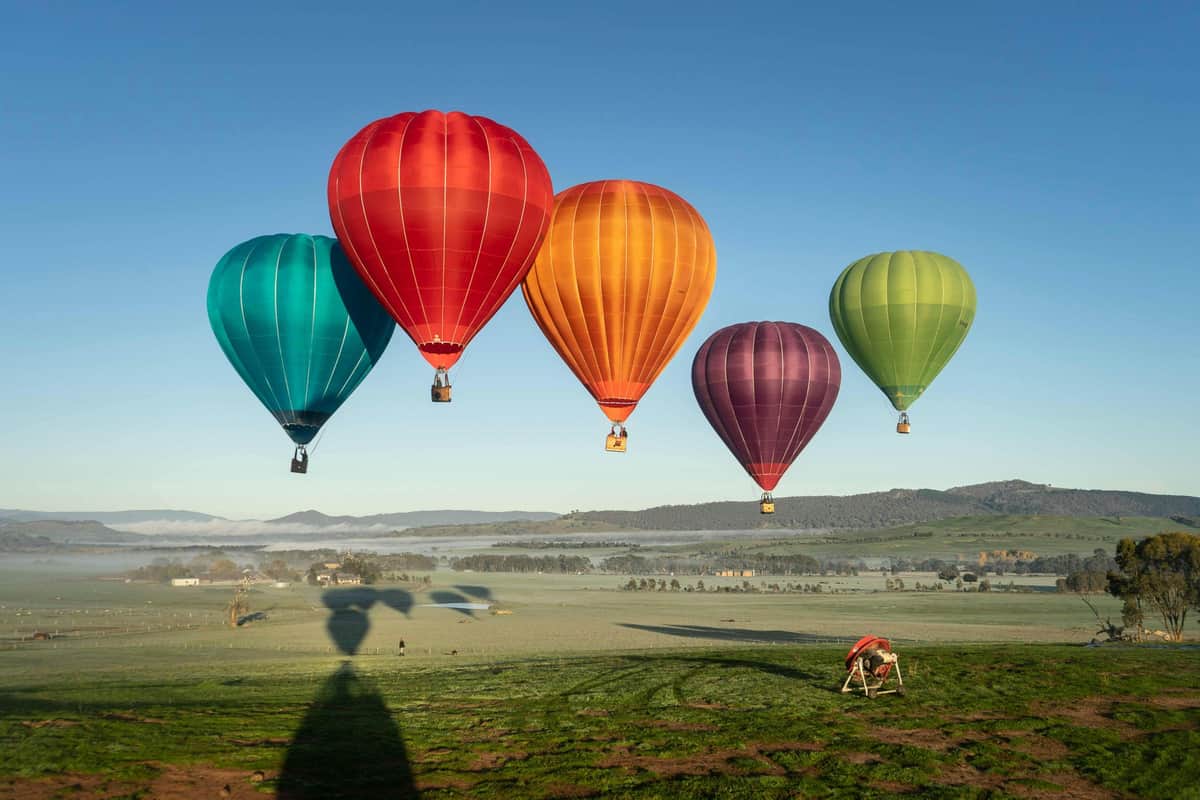 Cinq montgolfières colorées flottant au-dessus d'un champ vert avec des collines et un ciel clair en arrière-plan.