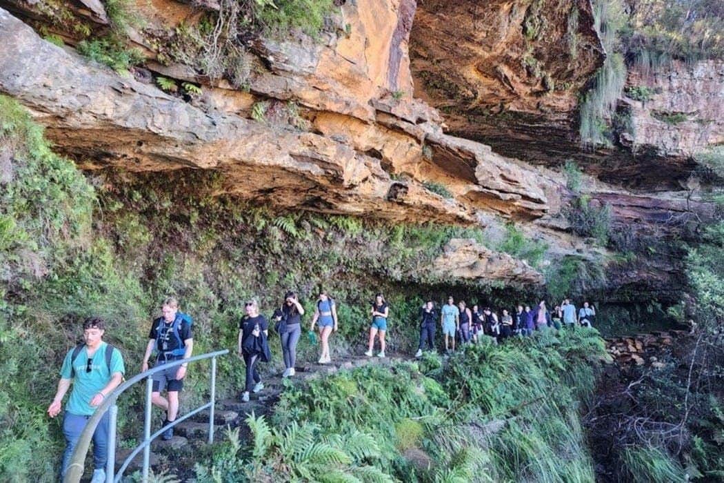 A group of people hiking along a narrow, rocky path lined with ferns and green vegetation under an overhanging rock formation.