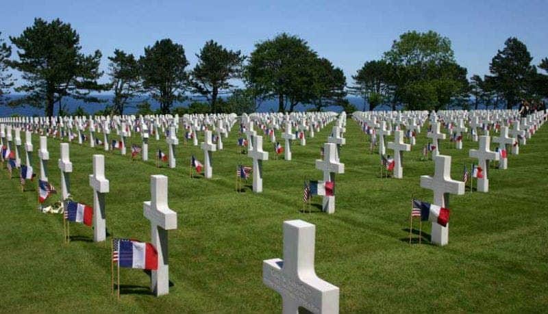 Reihen von weißen Kreuzen auf einem Friedhof mit französischer und amerikanischer Flagge, umgeben von Bäumen und unter einem klaren blauen Himmel.