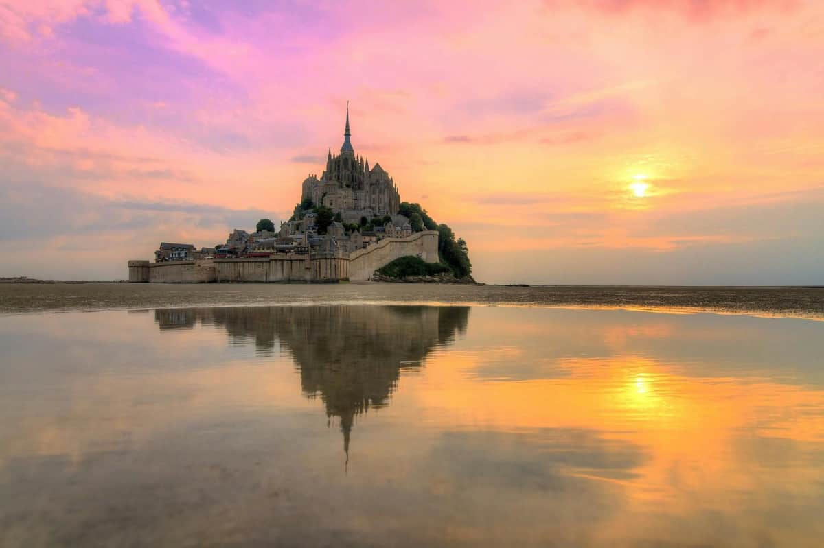 Mont Saint-Michel & Omaha Beach: Day Trip from Paris A castle-like structure on an island surrounded by water, reflecting in the foreground, under a colorful sunset sky.