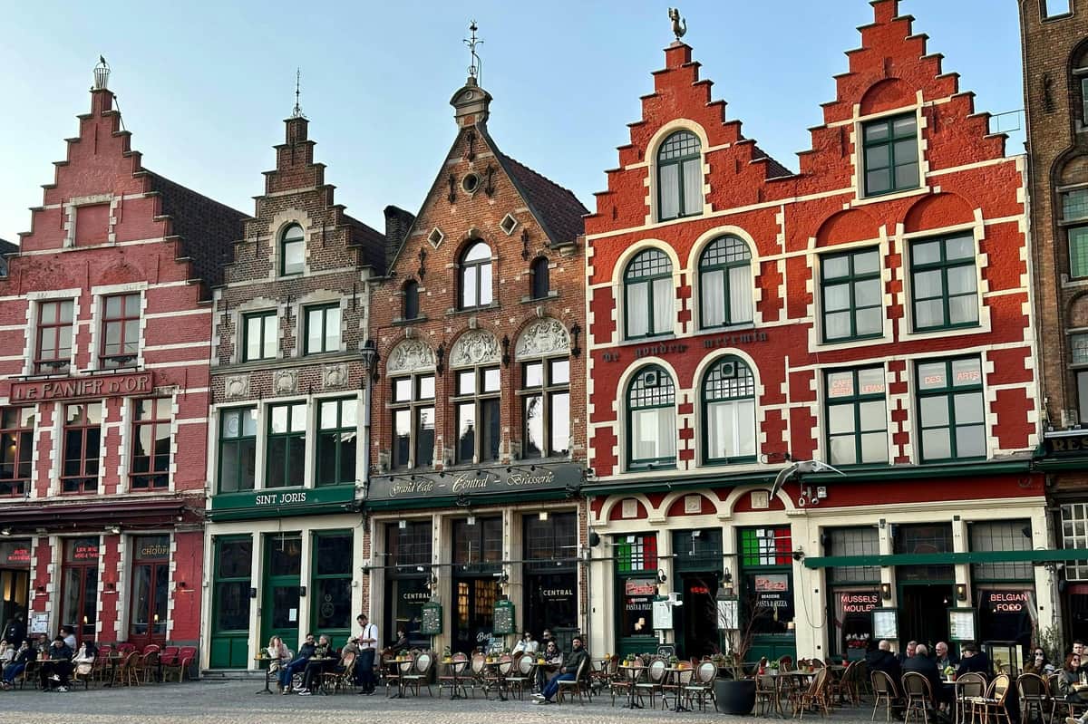 La gente se sienta en mesas de cafetería al aire libre frente a tres edificios históricos con fachadas ornamentadas y grandes ventanales.