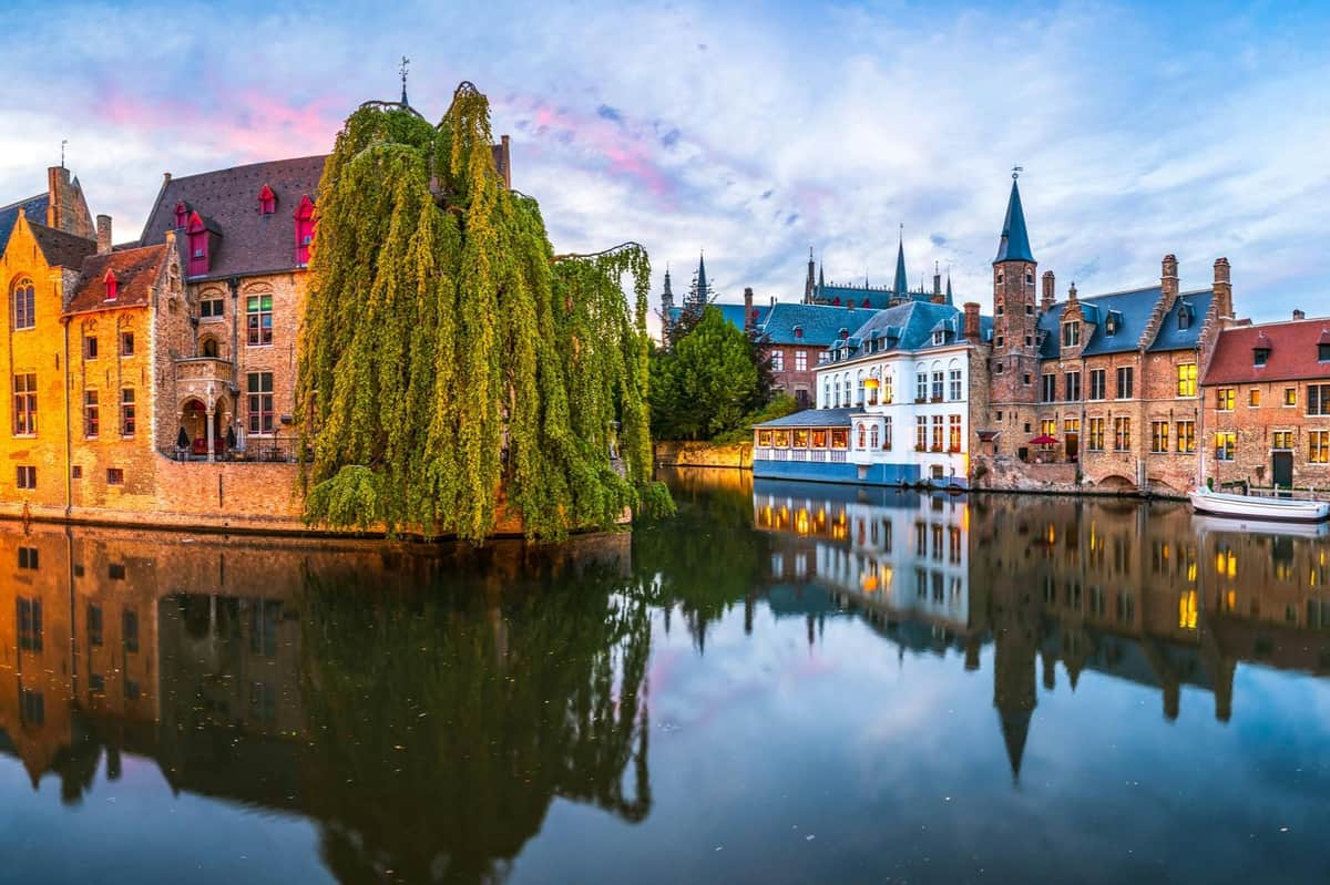 Bruges: Guided Tour from Paris A canal lined with historic buildings and a tree with hanging branches, reflecting in the water under a blue sky with pink clouds.