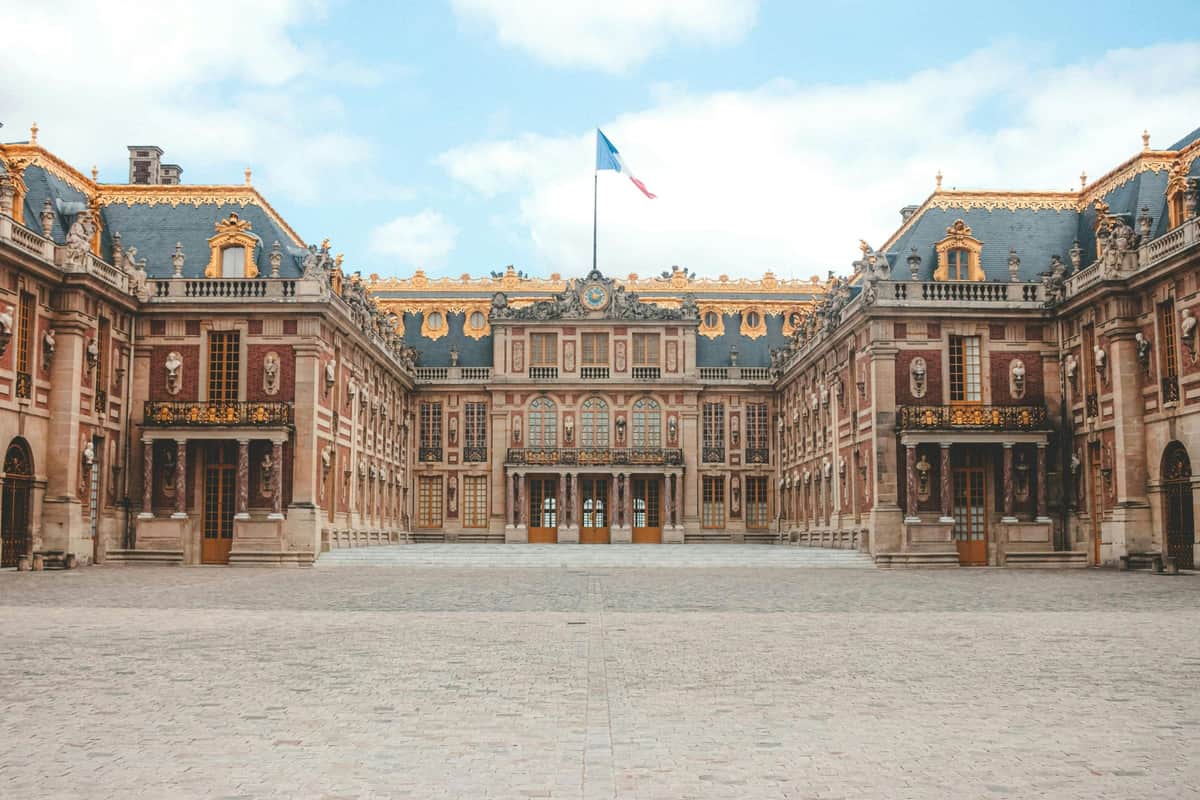 Eiffel Tower & Palace of Versailles: Small Group Guided Tour + Roundtrip Train A grand palace with ornate architecture, gold accents, and a French flag atop, viewed from a cobblestone courtyard.