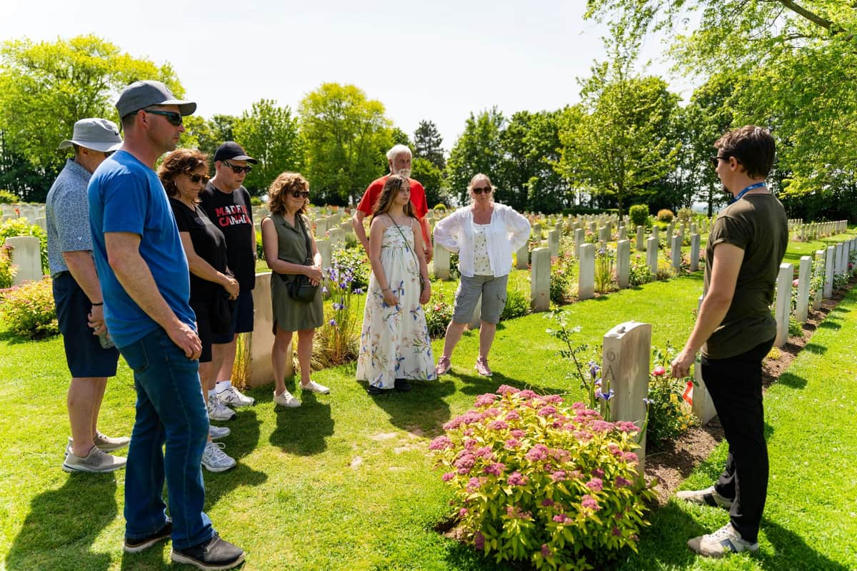 A group of people standing outdoors in a cemetery, surrounded by gravestones and greenery on a sunny day.