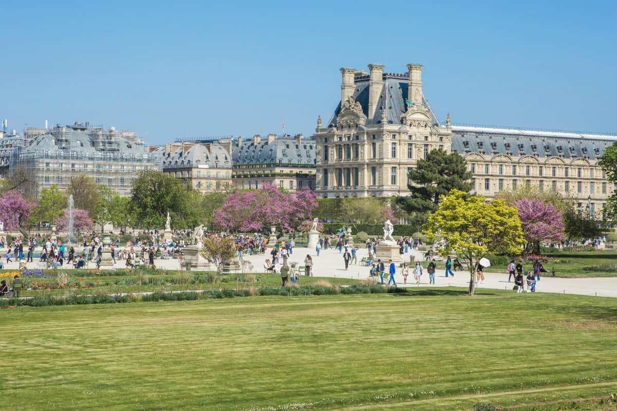People walking in a garden with trees, statues, and a historic building in the background under a clear blue sky.