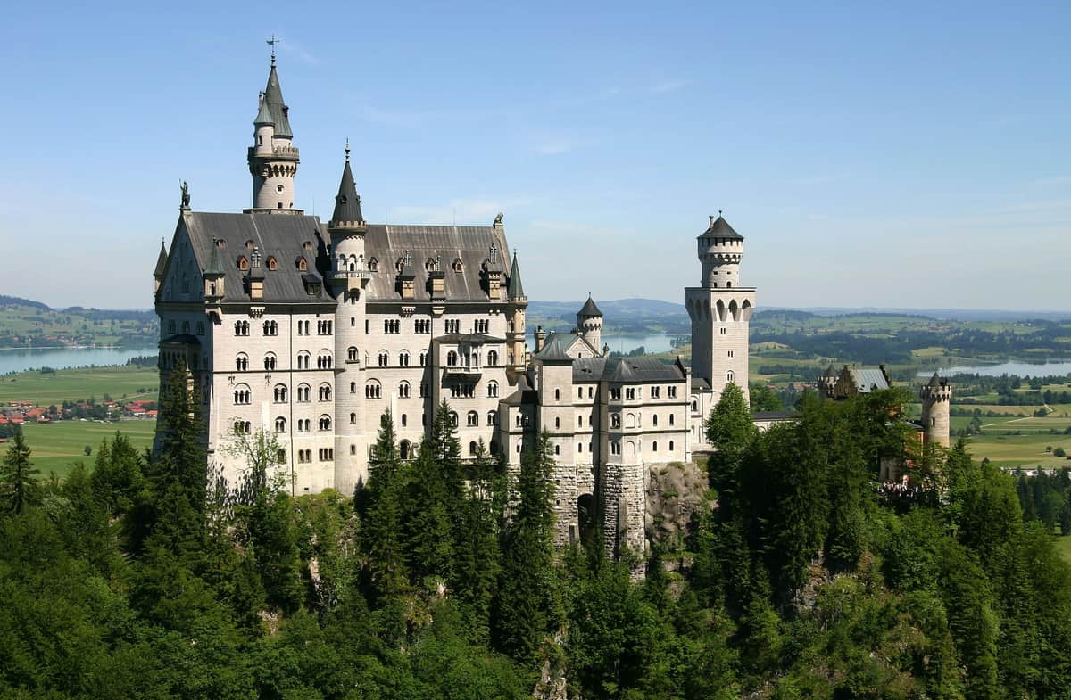 Ein großes, verziertes Schloss mit mehreren Türmen und Türmchen, umgeben von üppigen grünen Bäumen, mit Fernblick auf einen See und die Landschaft.