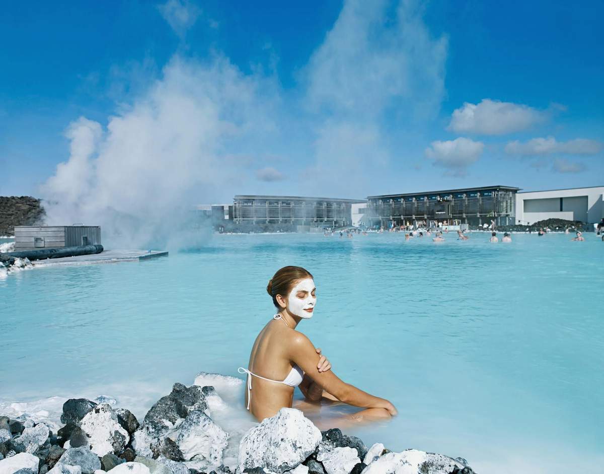 Blaue Lagune: Eintritt + Tagestour von Reykjavik (Reykjavík) A woman with a facial mask sits on rocks beside a blue geothermal pool, with buildings and steam in the background.