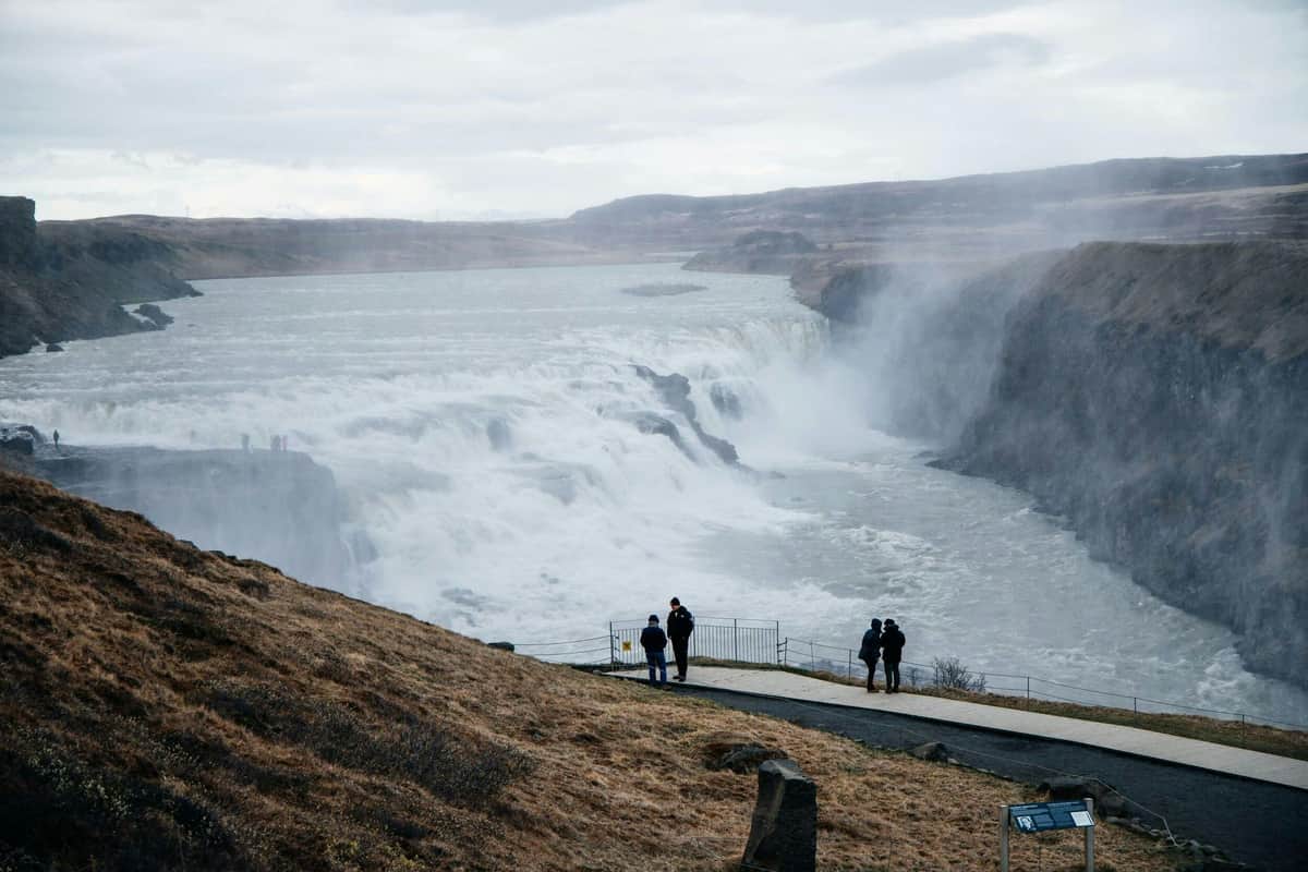 A group of people stand near a railing overlooking a large waterfall in a rugged landscape under a cloudy sky.