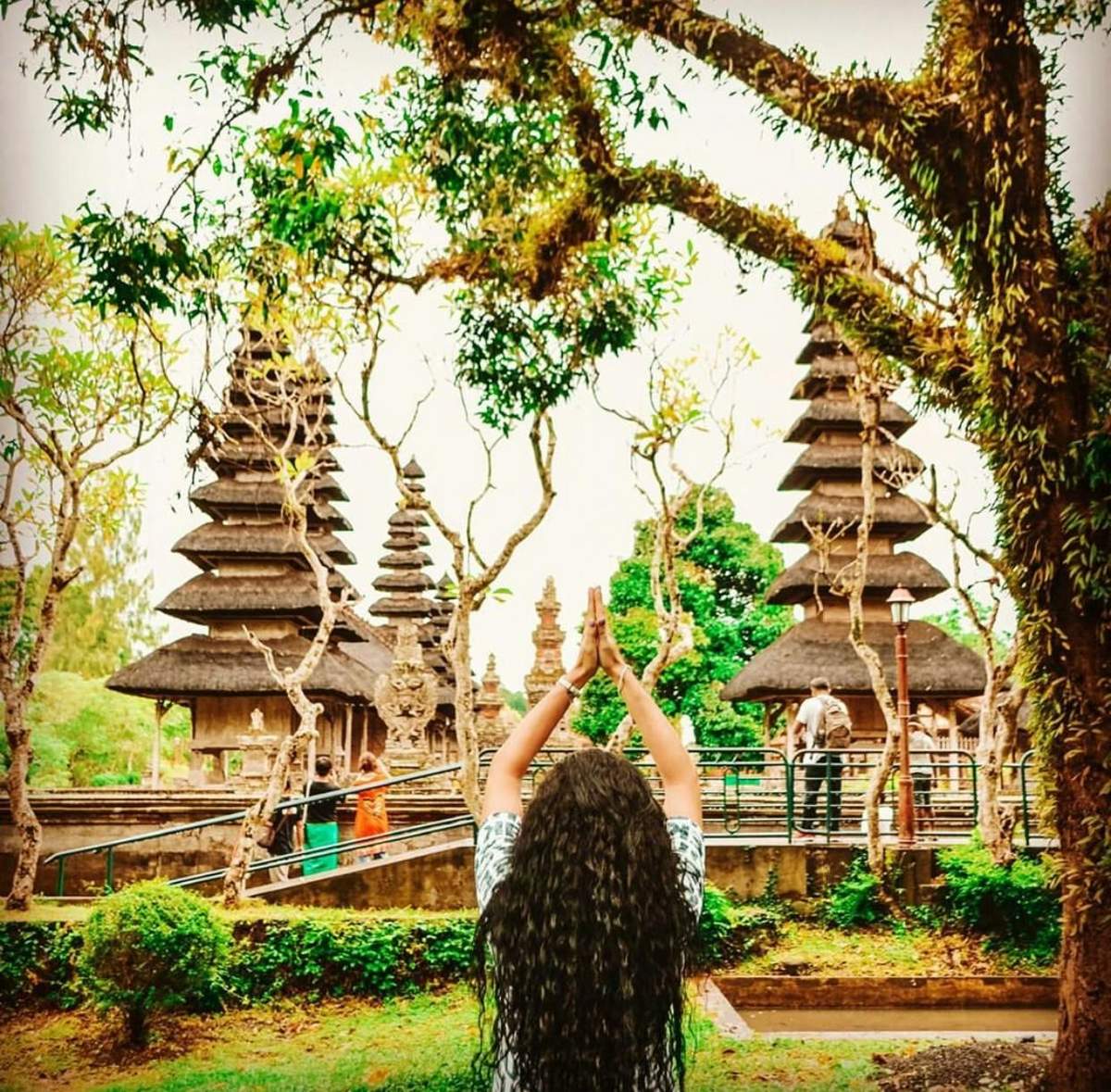 A person with long, curly hair raising their arms in front of traditional tiered towers in a lush, green temple garden.