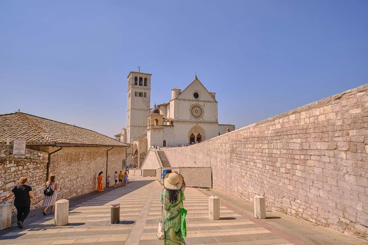 Una persona vestida de verde y con sombrero camina hacia una iglesia histórica con campanario bajo un cielo azul despejado.