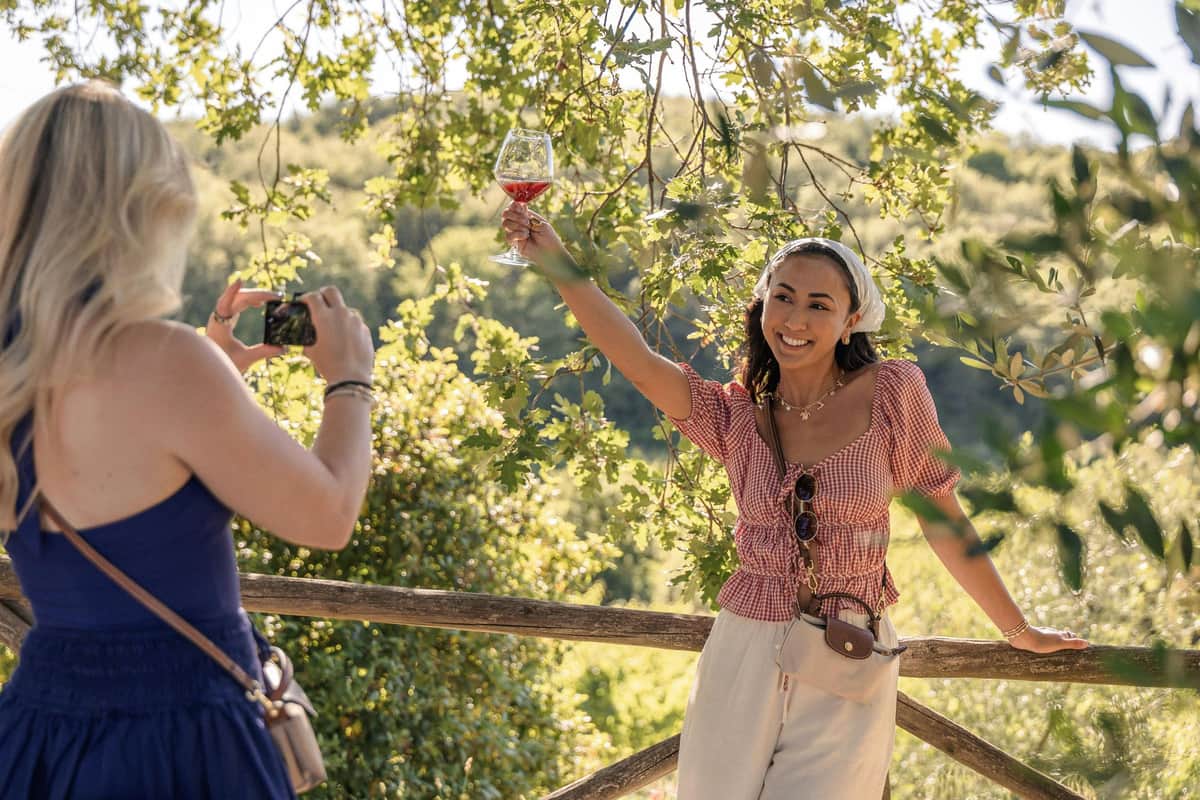 Una mujer con una copa de vino tinto en la mano, de pie al aire libre junto a una valla de madera, rodeada de árboles verdes y luz del sol.