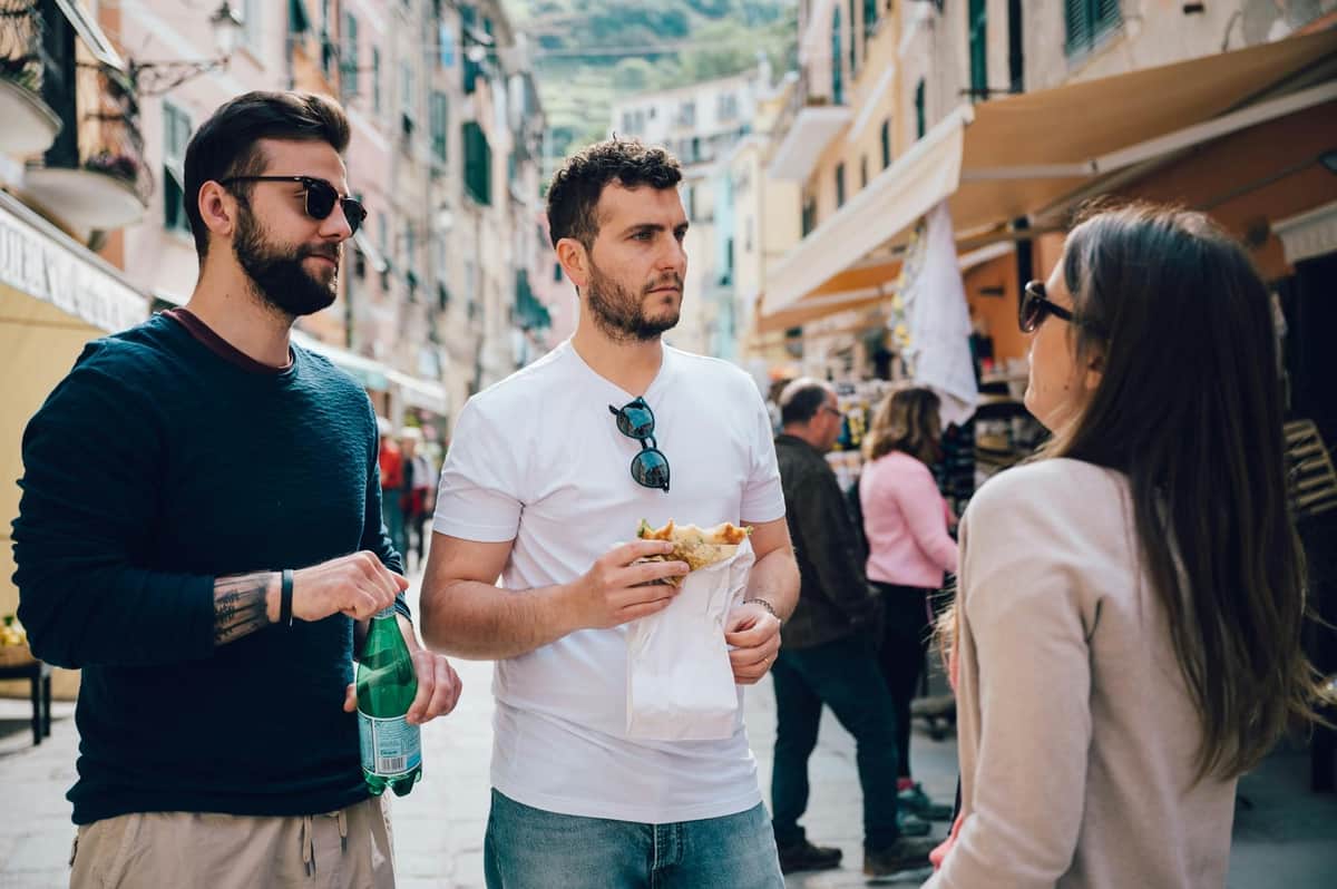 Two men in casual clothing stand on a bustling street. One holds a drink and the other holds food. People and buildings in the background.