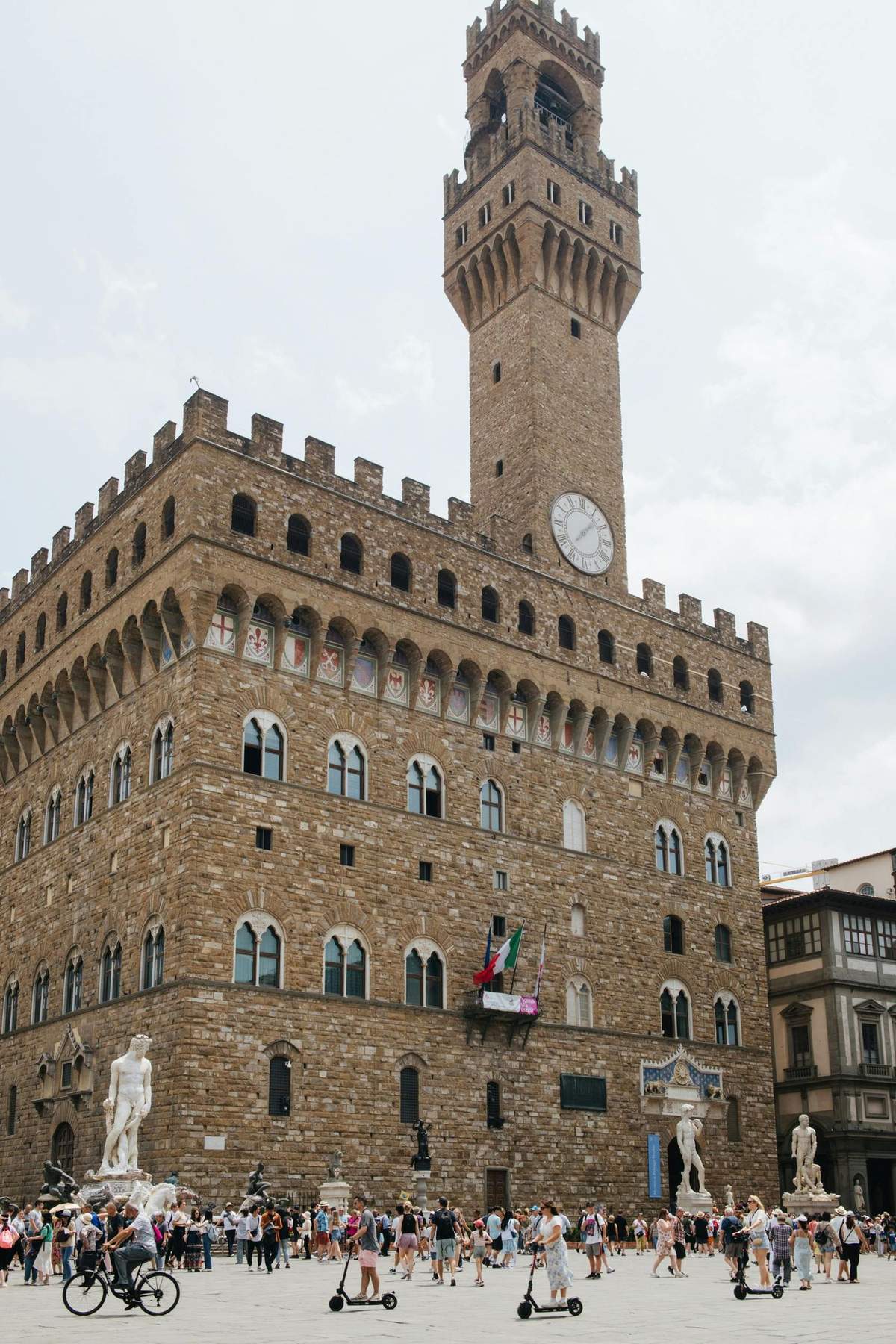 Un gran edificio histórico de piedra con ventanas arqueadas, una torre con reloj y una estatua delante. De un balcón cuelgan banderas italianas.