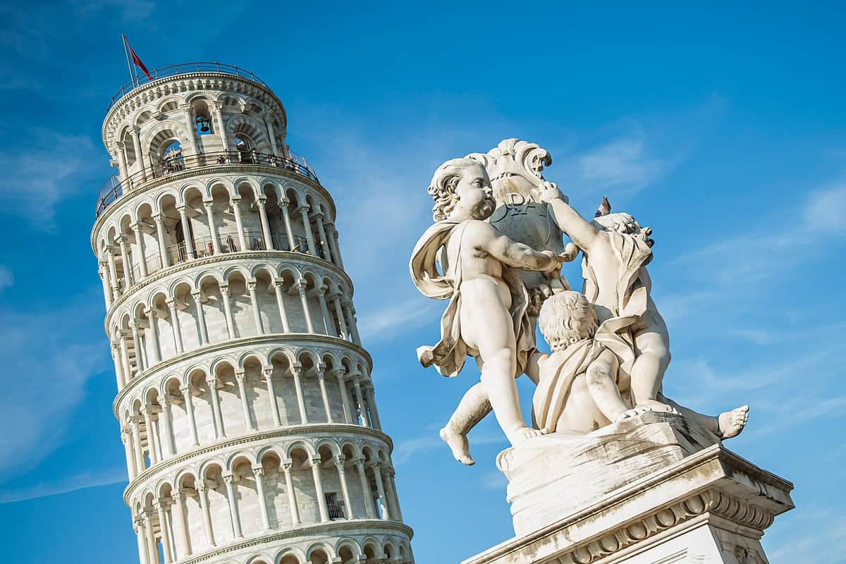 Estatua de mármol blanco de tres querubines frente a la Torre Inclinada de Pisa bajo un cielo azul despejado.