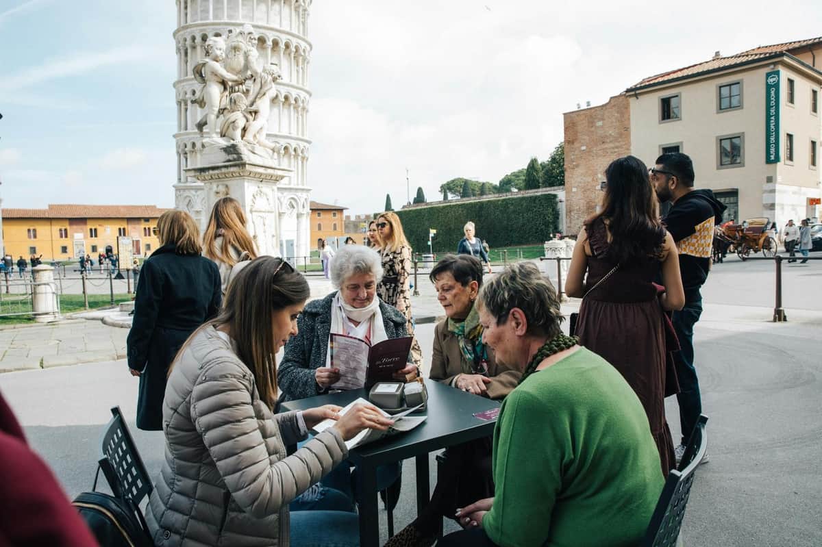 People sitting at an outdoor café table near the Leaning Tower of Pisa, with others walking and interacting in the background.