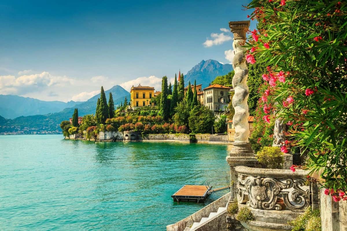 A scenic lakeside view with colorful buildings, lush greenery, and distant mountains under a clear blue sky.