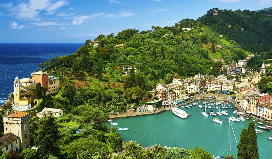 Une ville côtière avec des bâtiments colorés, des yachts amarrés dans un port turquoise, entourée de collines verdoyantes et d'un ciel bleu.