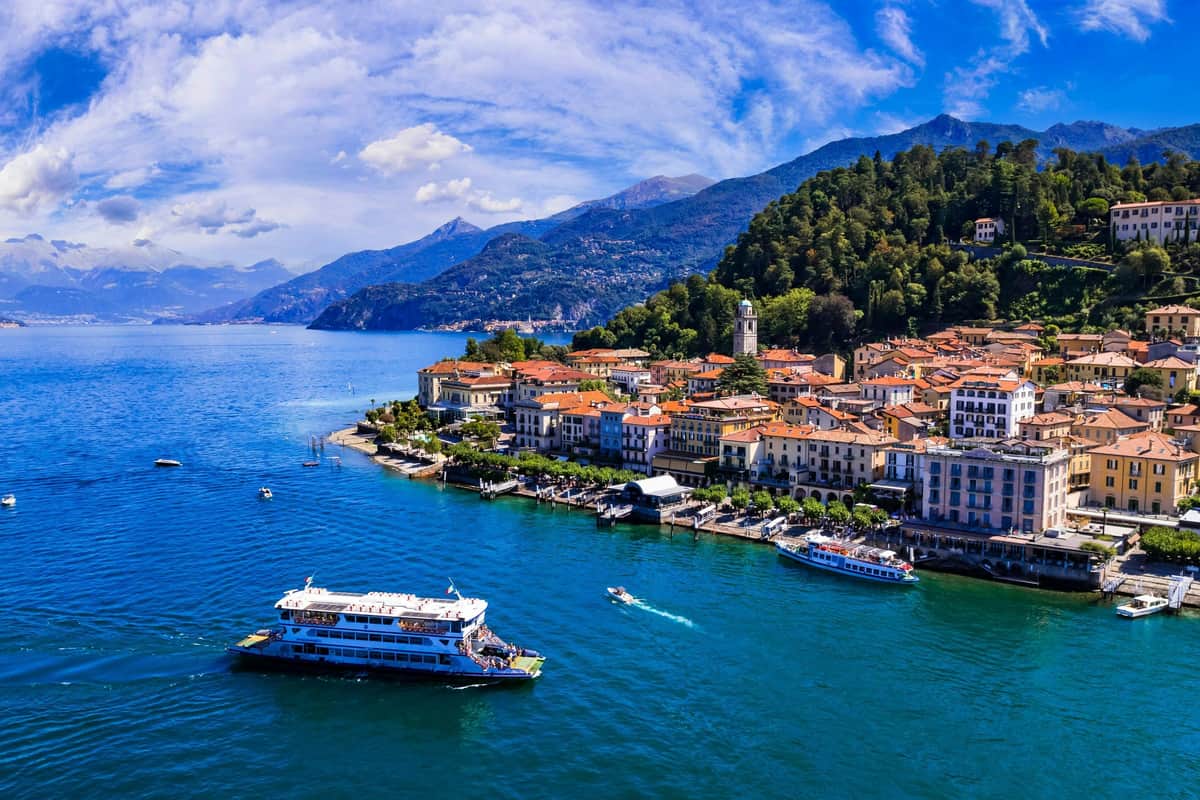 A scenic view of a lakeside town with colorful buildings, boats in the water, and mountains in the background under a partly cloudy sky.