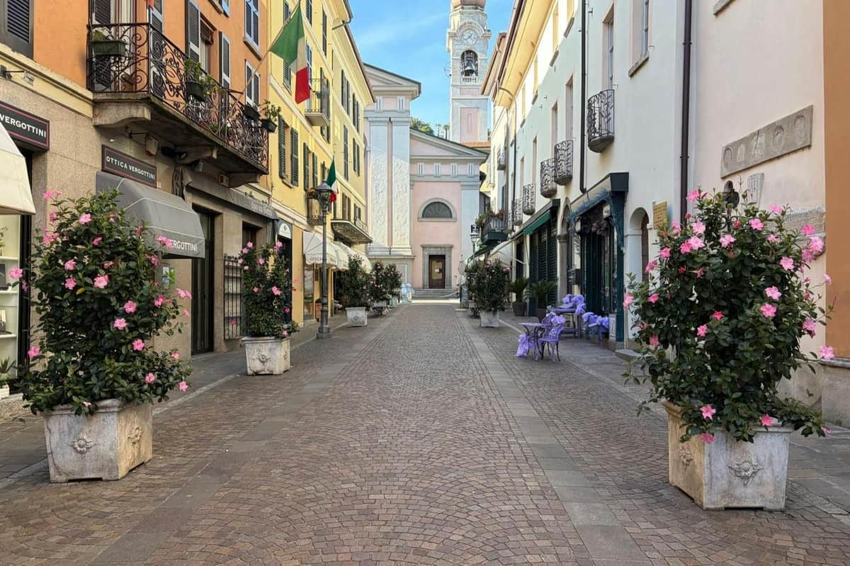 Lake Como, Bellagio & Lugano: Guided Tour from Milan + Ferry Ride A cobblestone street lined with buildings and potted plants leads to a church with a tall bell tower in the background.