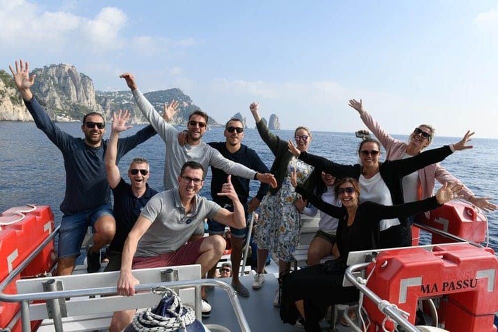 Eine Gruppe von Menschen, die lächelnd und mit erhobenen Armen auf einem Boot posieren, mit felsigen Klippen und dem Meer im Hintergrund.