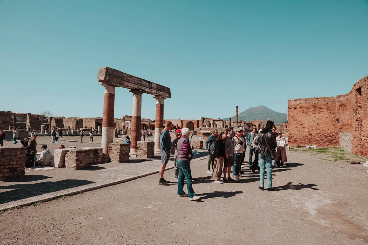 A group of tourists stands near ancient ruins with columns under a clear sky, with a distant mountain in the background.