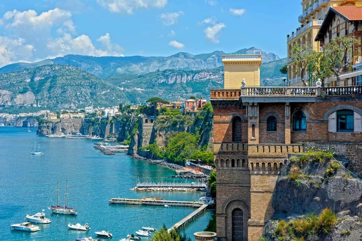 A scenic coastal view with cliffs, buildings, a marina, and mountains in the background under a partly cloudy sky.