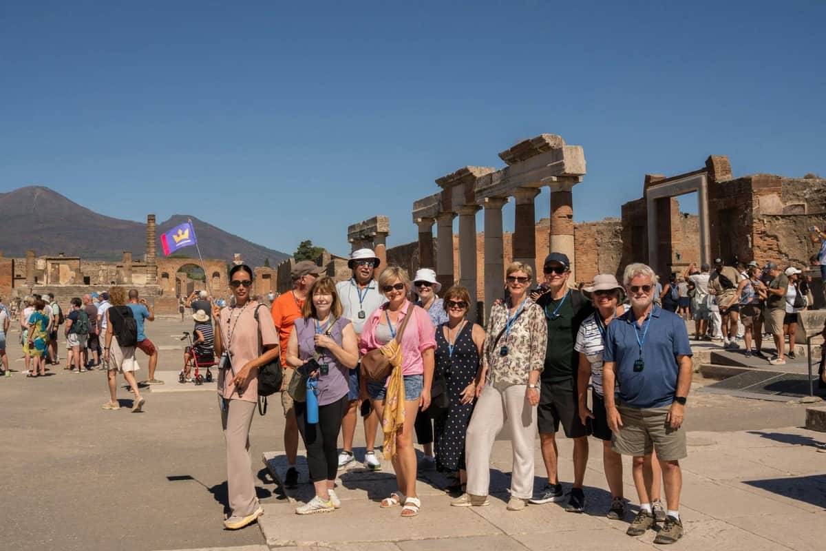 Eine Gruppe von Touristen vor antiken Ruinen mit Säulen, unter einem klaren blauen Himmel. Im Hintergrund sind weitere Touristen zu sehen.
