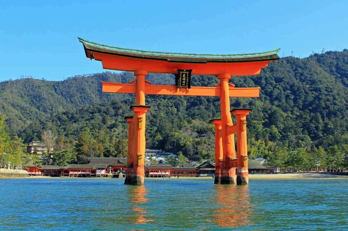 大きな赤い鳥居が水の中に立っており、裏には神社と森の丘が見えます。