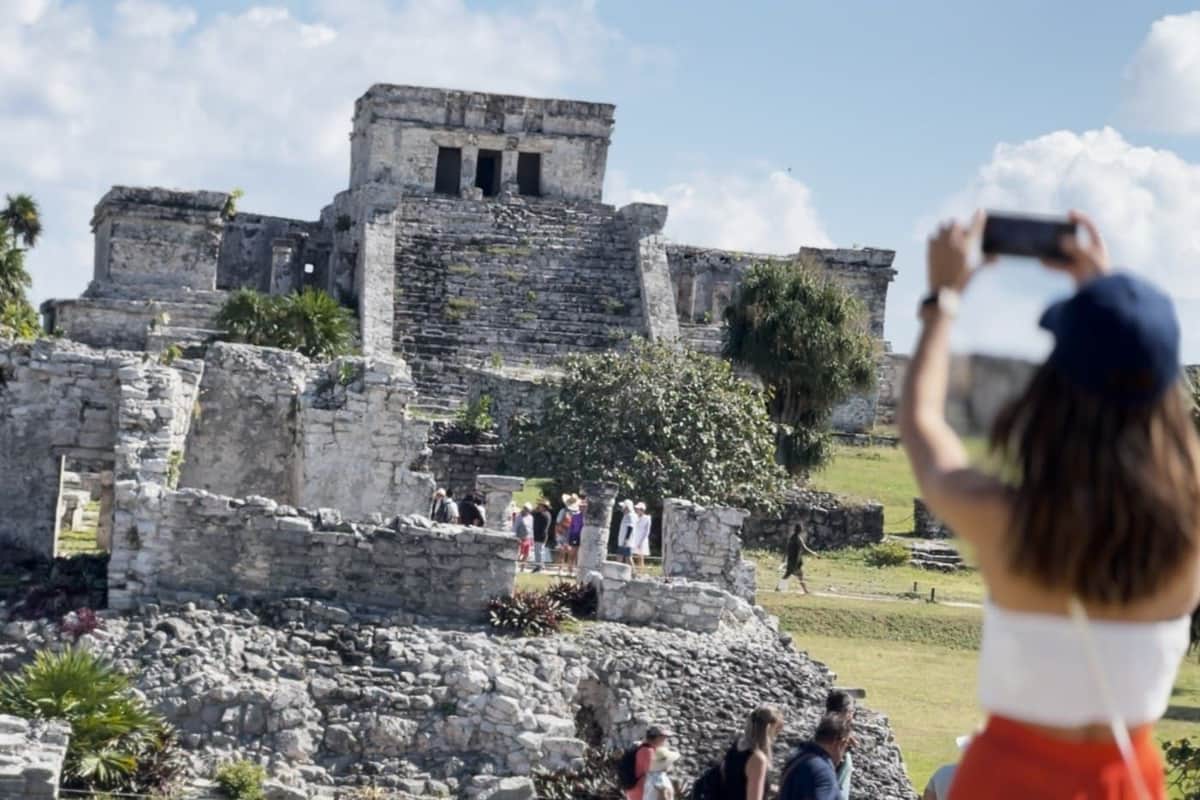 Ruinas Mayas de Tulum, Cenote Mariposa y Playa del Carmen: Viaje de ida y vuelta con almuerzo Turistas exploran unas antiguas ruinas de piedra bajo un cielo despejado, con una persona haciendo una foto en primer plano.