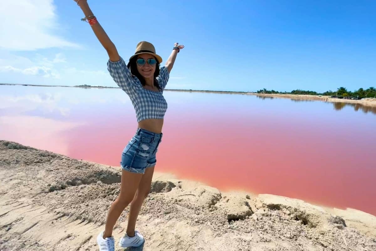 Une femme coiffée d'un chapeau et portant des lunettes de soleil, vêtue d'un haut à carreaux bleus et d'un short en jean, se tient debout, les bras levés, à côté d'un lac rose.