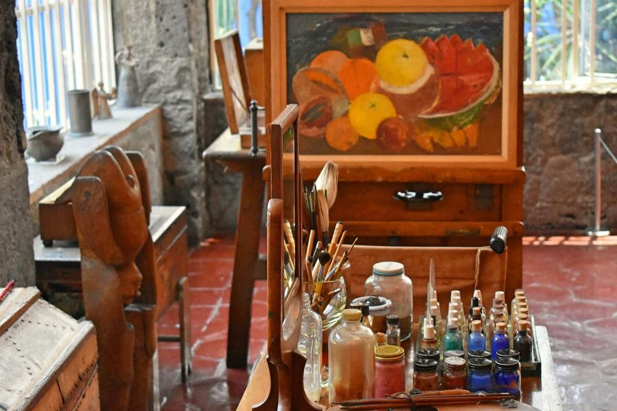 Xochimilco et Coyoacán : Visite guidée d'une journée + entrée au musée Frida Kahlo + transferts Un atelier d'artiste avec des bouteilles de peinture, des pinceaux et une palette. Une peinture de fruits colorée est exposée sur un chevalet à l'arrière-plan.