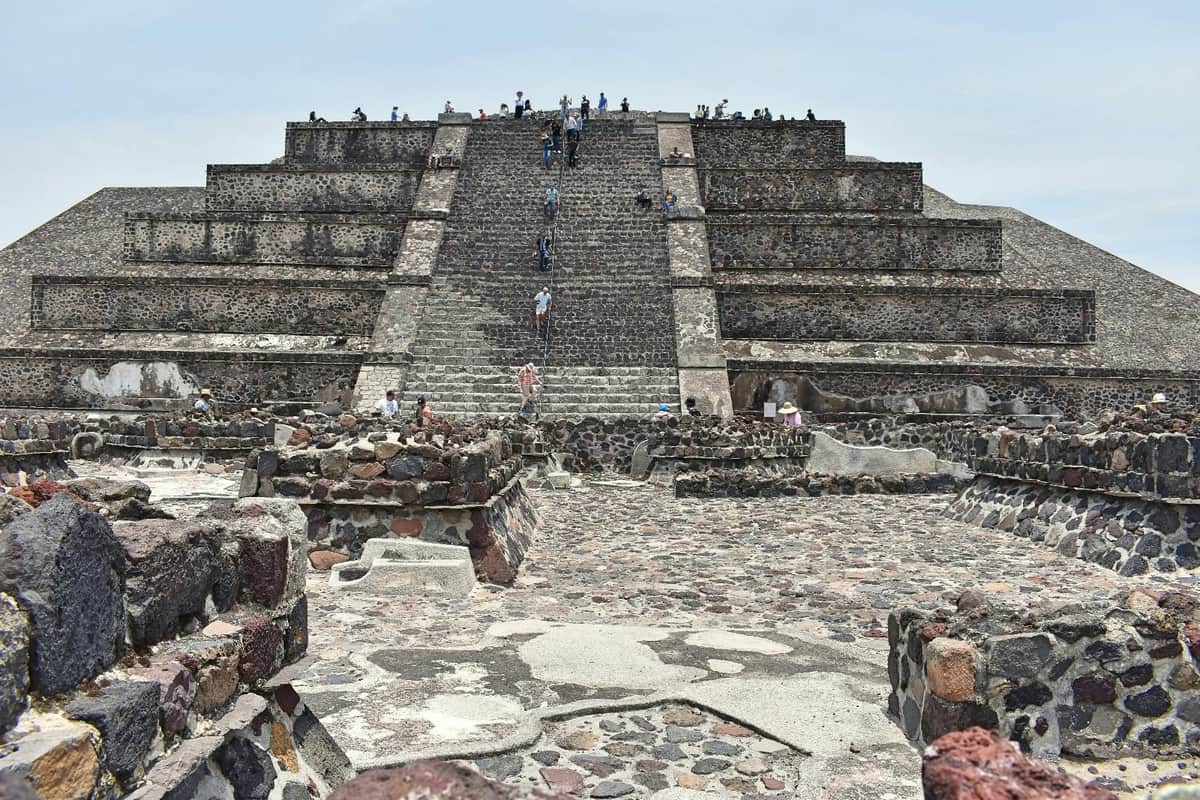 People climb the large, ancient stone pyramid steps at Teotihuacan, surrounded by ruins under a clear sky.