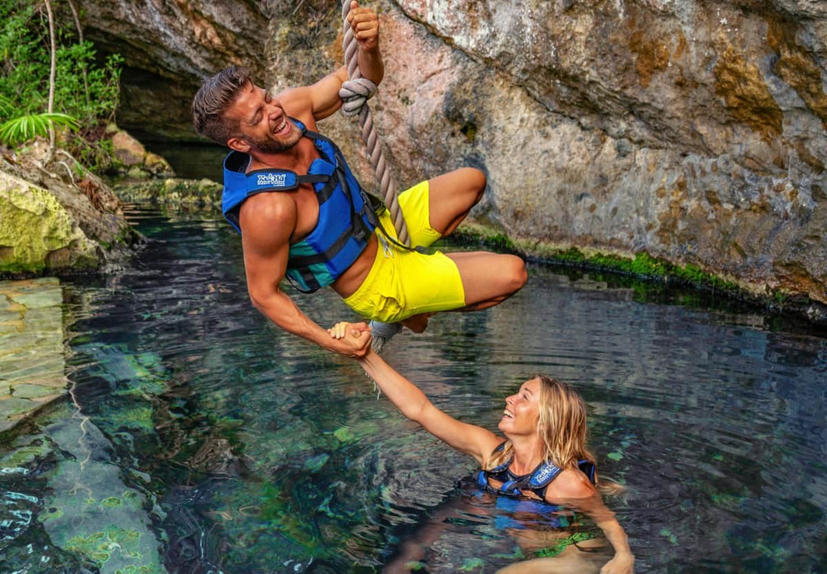 Man in yellow shorts and woman in blue swimsuit wearing life vests, smiling, and playing on a rope swing over rocky water.