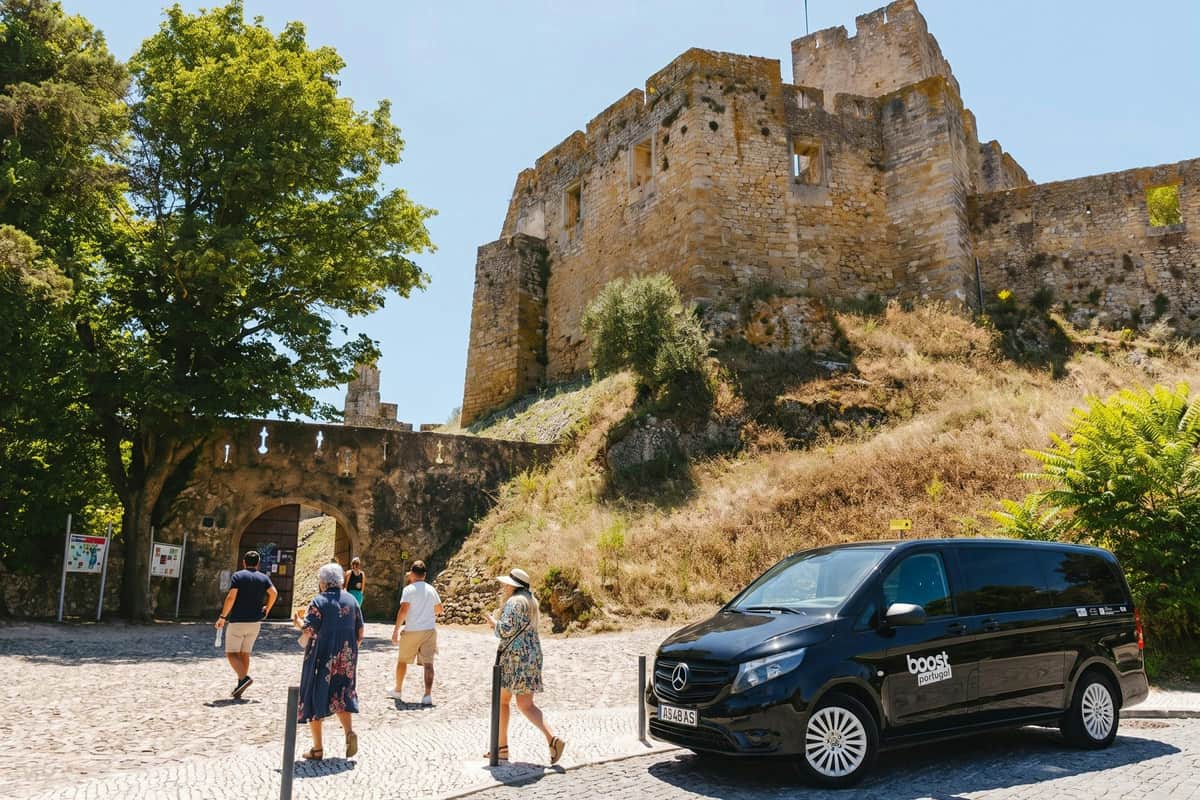 Des personnes se promenant près d'un château historique en pierre sous un ciel dégagé, avec une camionnette noire garée au premier plan.