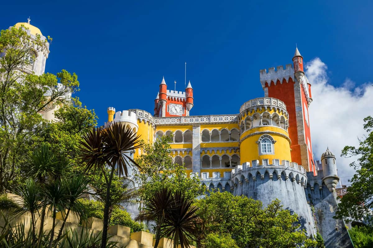 Palais de Pena, Nazaré et Fátima : visite guidée en petit groupe au départ de Lisbonne Un château aux couleurs vives, aux murs jaunes, rouges et blancs, entouré d'arbres sous un ciel bleu limpide.