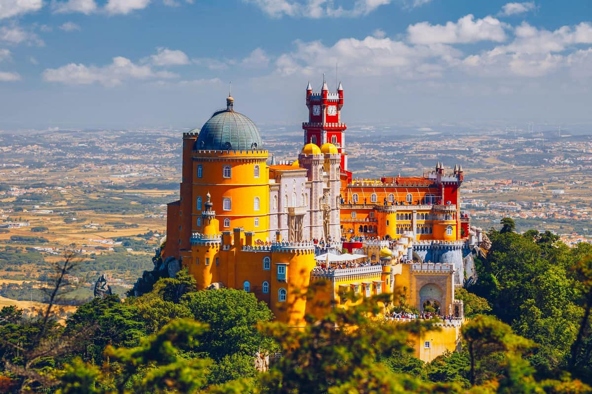 A colorful castle with yellow, red, and white sections on a hilltop surrounded by greenery under a blue sky.