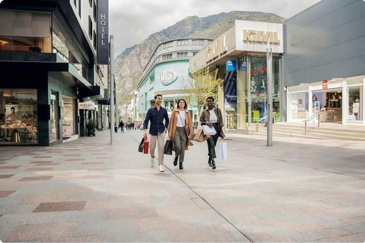 Trois personnes marchant dans une rue commerçante, des sacs de courses à la main, avec des immeubles modernes et des montagnes en arrière-plan.