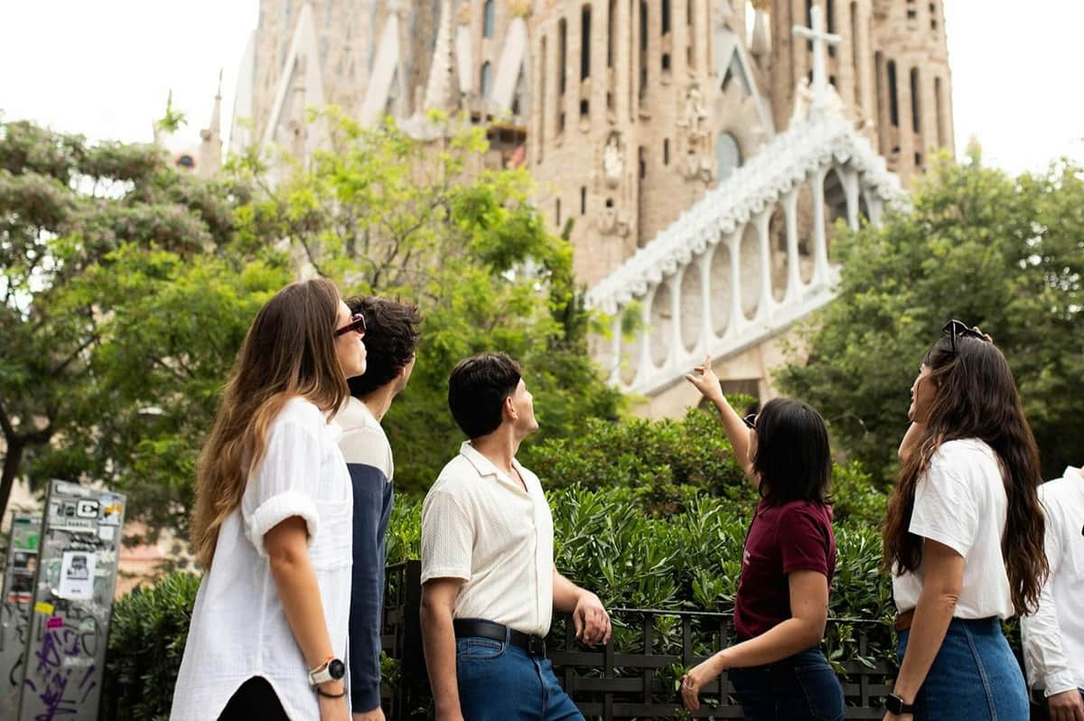 Trois personnes se tiennent dans un parc, le regard tourné vers la Sagrada Familia en arrière-plan, au milieu de la verdure et d'éléments urbains.