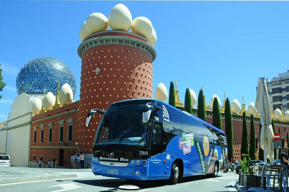 Bus pour Figueres : Carte journalière depuis Barcelone ou Gérone Un bus bleu portant l'inscription "Barcelona Bus" est garé devant un bâtiment rouge unique dont le toit est orné de grandes sculptures en forme d'œuf.