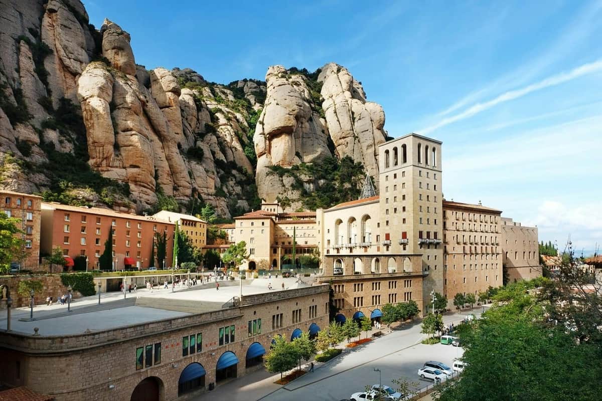 Ensemble de bâtiments historiques avec des arches et une haute tour au pied de montagnes rocheuses et escarpées, sous un ciel bleu et clair.