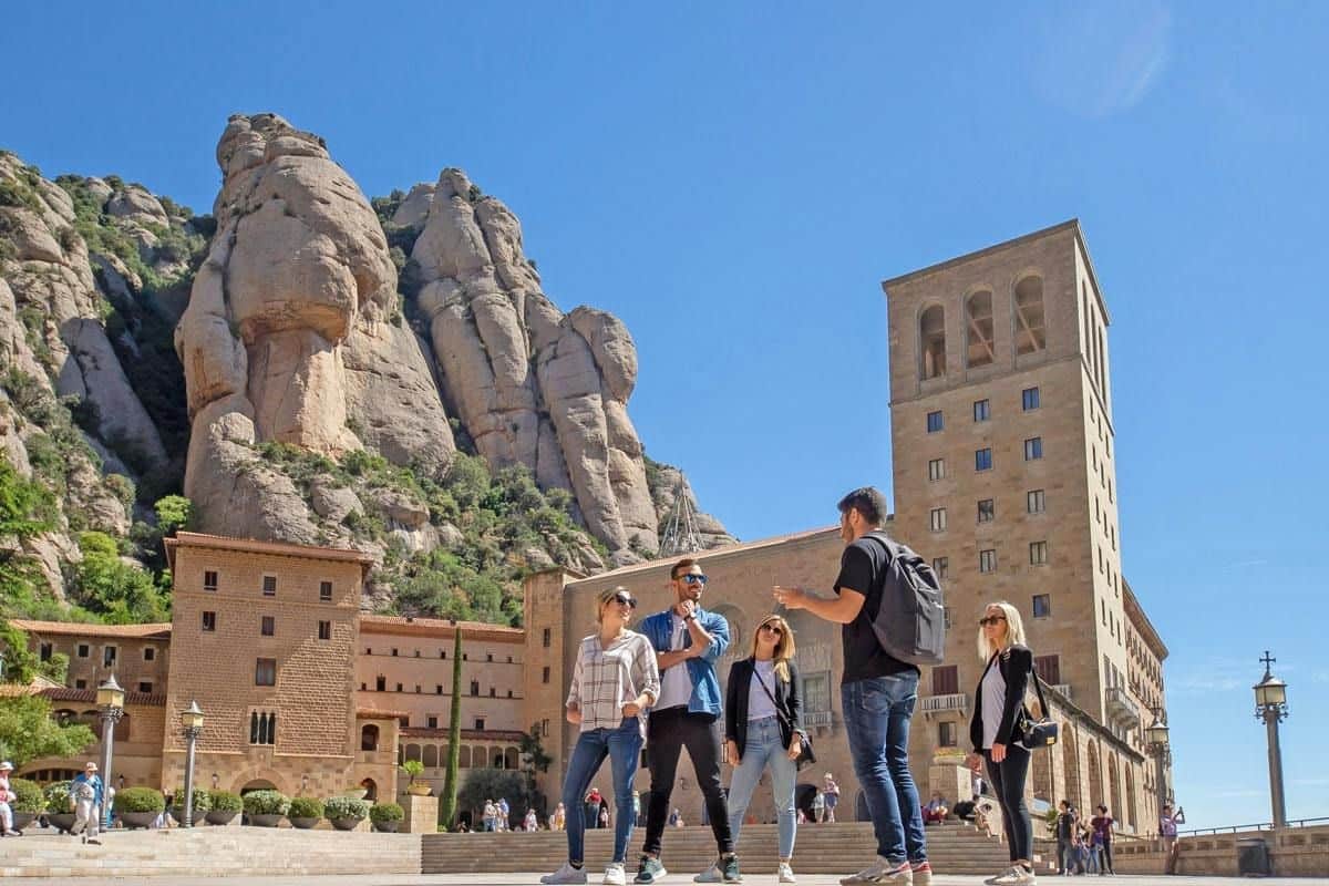 Un groupe de personnes debout, en train de discuter sur une place située près d'un bâtiment historique et de falaises, sous un ciel bleu limpide.