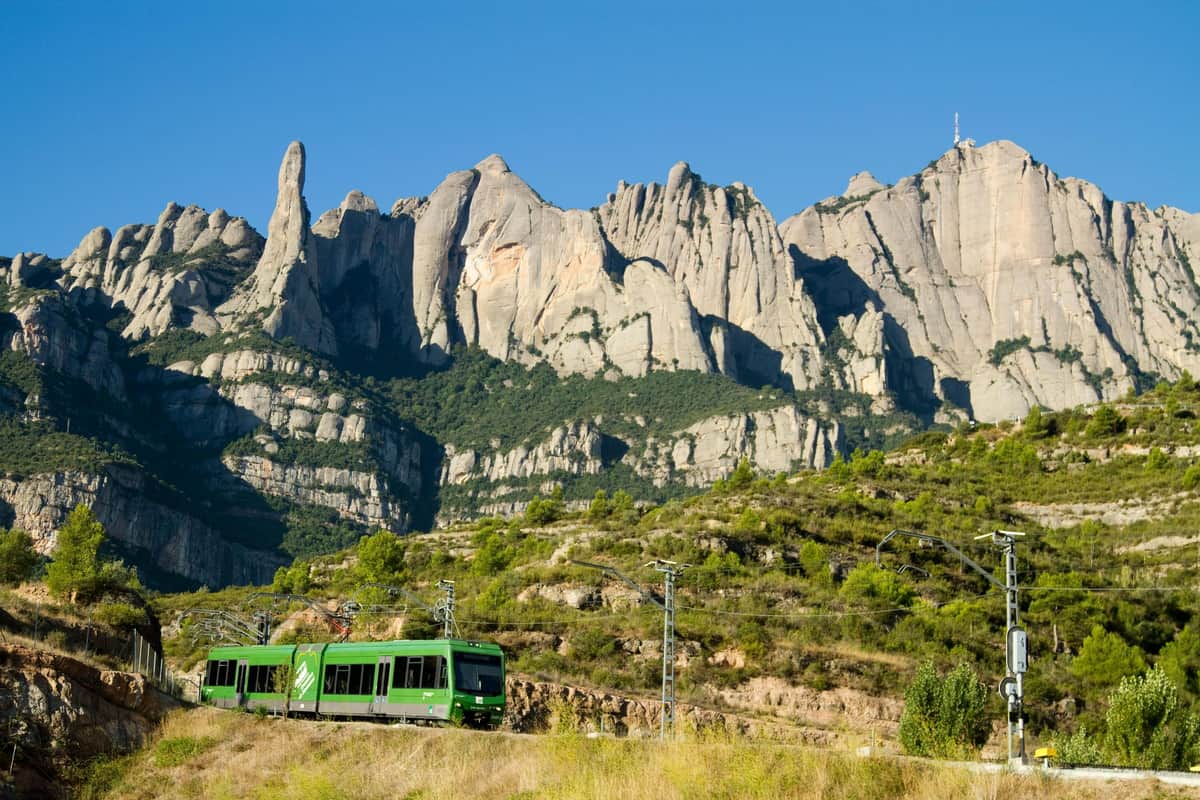 A green train travels through a hilly area with rugged, rocky mountains in the background under a clear blue sky.
