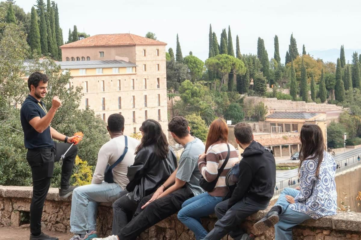 Montserrat : visite guidée d'une demi-journée depuis Barcelone Un guide touristique fait des gestes enthousiastes tout en s'adressant à un groupe de personnes assises en plein air, avec un bâtiment et des arbres en arrière-plan.