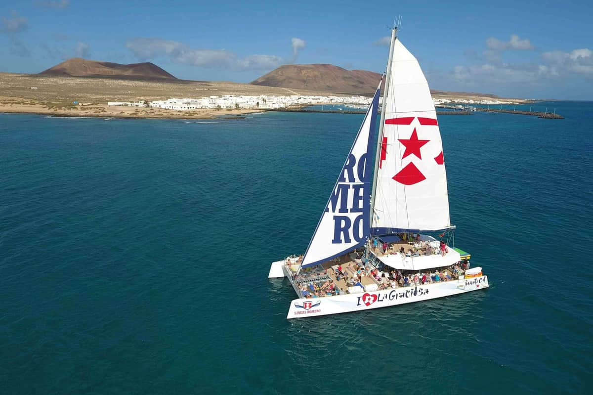 Catamaran with "I ❤️ La Graciosa" and star on sails, sailing in blue water near a coastal town with white buildings and hills.