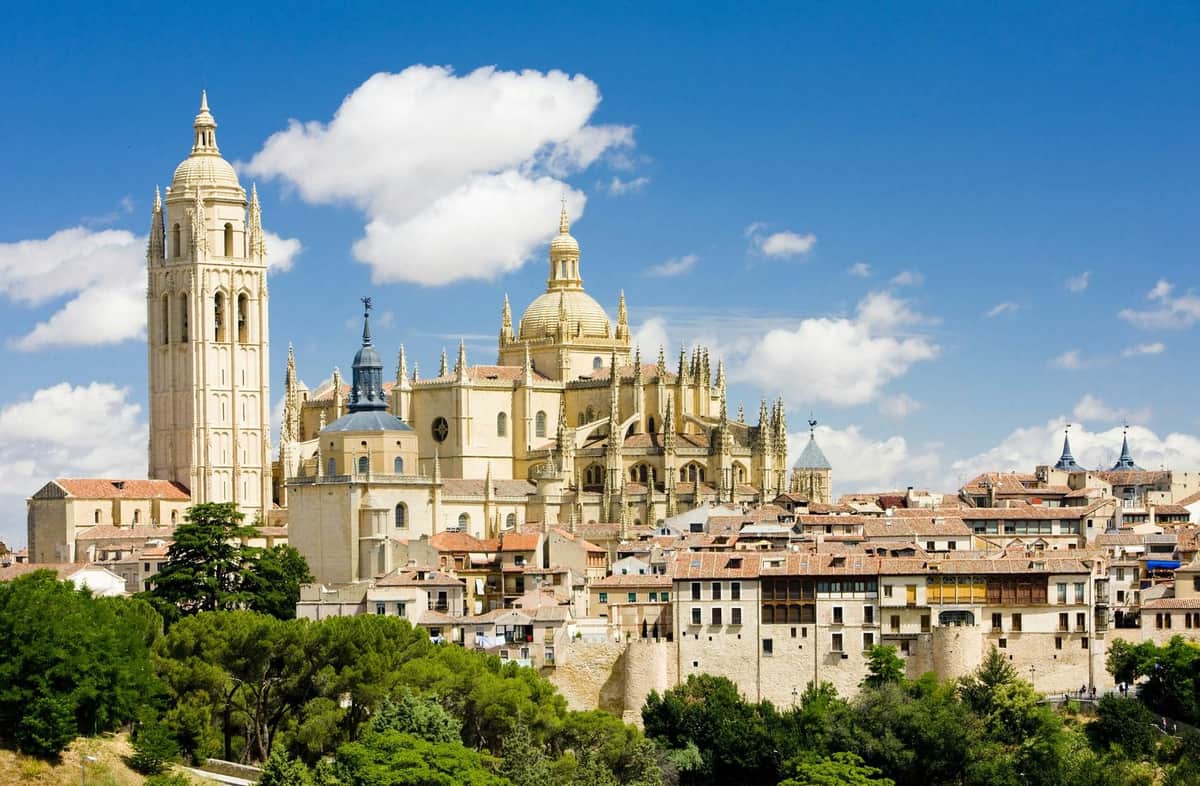 View of a Gothic cathedral with spires and large central dome, surrounded by historic buildings and lush greenery under a blue sky.
