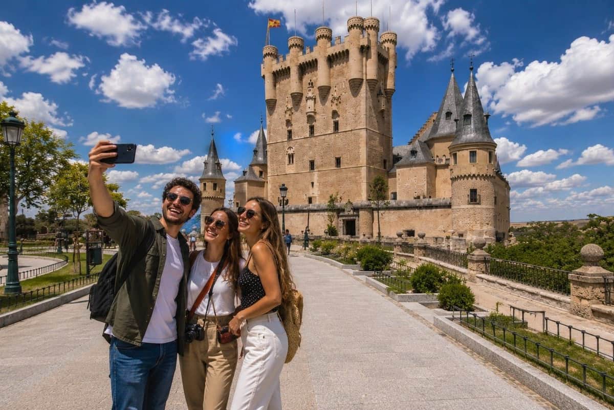 Segovia & Avila: Guided Walking Tour from Madrid Three people smile for a selfie in front of a historic castle under a sunny, cloudy sky.