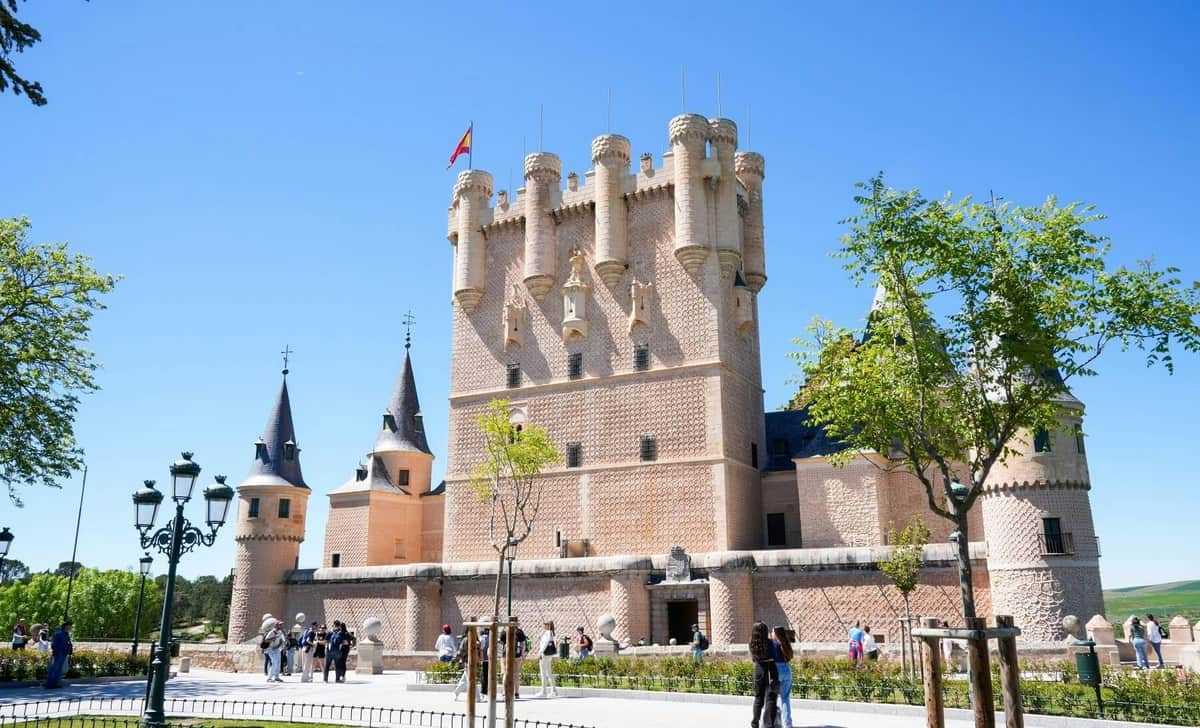 A medieval castle with a flag on top, surrounded by tourists, trees, and a clear blue sky.