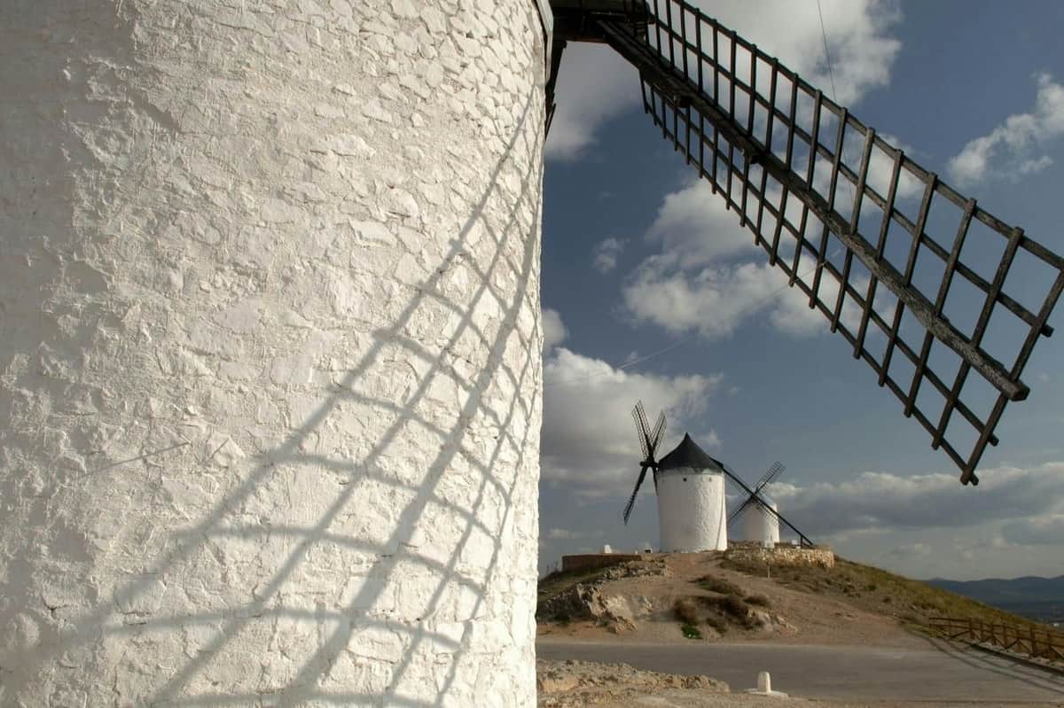 Close-up of a white windmill with its shadow on the wall, with two other windmills in the background under a cloudy sky.